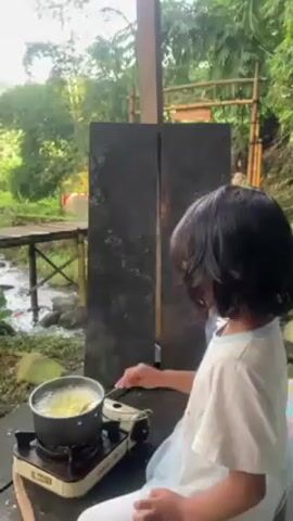 A young person with long dark hair is cooking something in a pot on a portable stove. The pot is silver and contains a yellow-colored substance that appears to be boiling. The person is using a spoon to stir the contents of the pot. The stove is sitting on a black surface, possibly a table or countertop.

The person is standing under a wooden structure with a partial roof. Behind them are two tall, dark gray panels. The background reveals a lush, green outdoor environment. A small stream flows nearby, crossed by a rustic wooden bridge. The scene is filled with greenery, including trees and various plants.