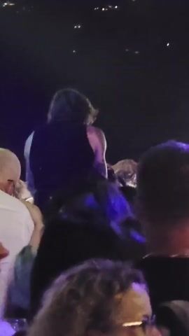 A short video shows a person singing into a microphone. The person has long, dark hair and is wearing a black tank top. They are on a stage with a dark background, and there are lights in the distance. There are people in the foreground, some of whom are out of focus. One person in the foreground is wearing a white shirt. The lighting is dim and colorful, with purple and pink lights visible. The person singing appears to be performing for an audience.