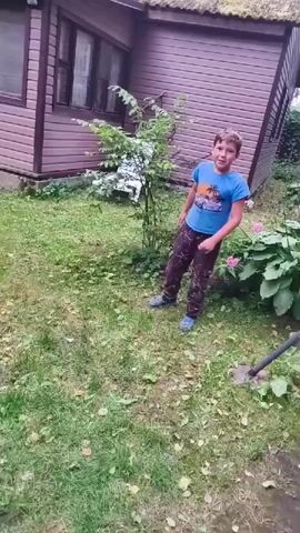A young boy in a blue t-shirt and camouflage pants runs across a grassy yard. He is in front of a wooden house with a thatched roof. The boy moves from left to right across the screen, with his body angled towards the camera. He appears to be in motion, possibly running or jumping. The yard is covered in green grass and some scattered fallen leaves. There is a small tree to the left of the boy and some pink flowers to his right. The camera appears to be following the boy's movement.