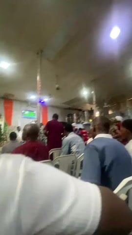 The video shows an indoor gathering of people. The camera is positioned behind a person in the foreground, with their white shirt sleeve visible on the left. The gathering appears to be in a hall or church with a suspended ceiling and lights. Several individuals are seated on white chairs, facing a stage or focal point at the front of the room. Some people are standing and walking around. On the left side of the room, there is a plant and some orange fabric draped on a wall. A green screen or display is visible on the stage area. The lighting is somewhat dim, with bright spotlights illuminating the ceiling. The people are diverse in age and gender, and many are dressed in casual or semi-formal attire. There is a sense of collective activity, with some individuals appearing to be engaged in conversation or attentively watching something at the front of the room.