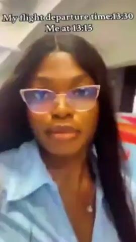 A video shows a woman walking through an airport. The text overlay states that her flight departs at 13:30 and she is there at 13:15. The camera is held up close to her face initially, showing her wearing glasses and a light blue collared shirt. She smiles briefly. The camera is then pointed downward, showing the tiled airport floor as she walks.  Sound is playing, with background music and possibly some ambient airport noise.