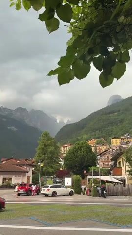 This is a video of a street scene in Molveno, Italy, with a focus on the dramatic mountain landscape in the background. The foreground shows a paved area with yellow parking lines, suggesting a parking lot or public square. A white station wagon is parked, and two people are visible near a red car. There's a building on the left with a sign that includes the word "Pyrex" and possibly "Gelateria" or a similar establishment. A sign on the right reads "GELATERIA" and "SNACK DRINK." A motorcycle is parked to the right of the Gelateria.

The mid-ground features more buildings, characteristic of a European town, nestled on the slopes of a densely forested mountain. The architecture suggests a tourist area, with colorful buildings and balconies. Lush green trees frame the scene, with a prominent branch of leaves hanging from the top of the frame, creating a natural border.

The background is dominated by majestic, rugged mountains shrouded in dramatic, cloudy skies. The clouds create an atmospheric and somewhat ominous mood, with glimpses of light breaking through the peaks. The lighting suggests it might be late afternoon or early evening, with overcast weather. The overall impression is a picturesque mountain village preparing for or experiencing a change in weather, with the grandeur of nature juxtaposed against the charming human settlement.