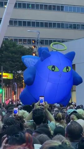 A large, inflatable blue cat with wings and a halo floats above a crowd of people. Many people in the crowd are holding up their phones to take pictures or videos of the inflatable cat. The scene appears to be outdoors, possibly in a city, with buildings and traffic lights visible in the background.