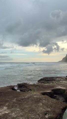 The scene is a rocky beach with ocean waves rolling in under a cloudy sky. The rocks in the foreground are dark and covered with some green vegetation. The ocean appears calm, with small waves breaking in the distance. A green cliff is visible at the right side of the frame. The lighting is soft due to the overcast sky. There are no people visible in the shot.