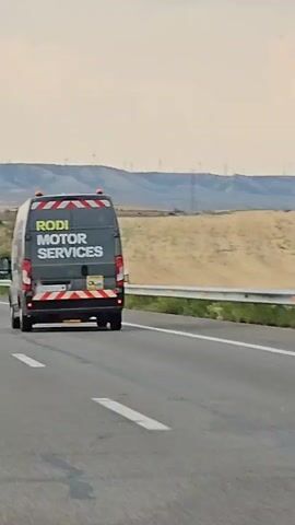 A dark grey van with yellow lettering that reads "RODI MOTOR SERVICES" is driving on a highway. The van has hazard lights on its roof and reflective tape on its back. The van's license plate is visible, showing "6220 LGM". The environment appears to be a rural area with rolling hills and wind turbines in the distance. The sky is overcast. The video is shaky, suggesting it was filmed from another moving vehicle.