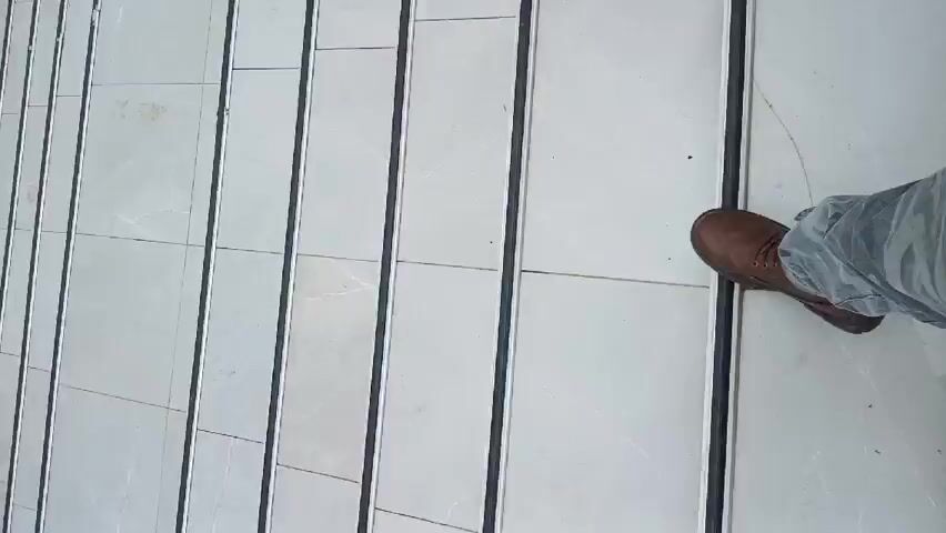 The video starts with a close-up of steps with white tiles and metal bars separating them. A person's foot, wearing a brown shoe and blue jeans, is visible on one of the steps. The camera then pans up, revealing a brick tower-like structure on a paved area. The background shows a landscape with greenery and a cloudy sky. A car can be seen parked in the distance.