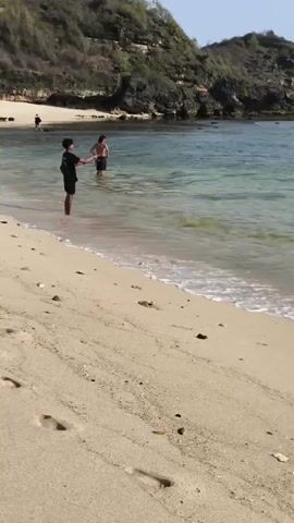 The video shows a person on a sandy beach with footprints visible. In the shallow water, two people are standing and interacting. The water is clear and shows gentle waves. In the background, there are rocky cliffs covered in vegetation, and the sky is clear and blue. The scene is a coastal environment with natural geological formations.