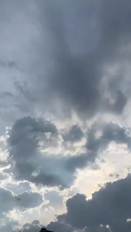 The video shows a view of clouds in the sky. The clouds are dark gray and heavy, with some lighter patches of blue sky visible between them. The sun appears to be behind the clouds, casting a diffused light. There is no discernible action or interaction taking place. The environment appears to be outdoors.