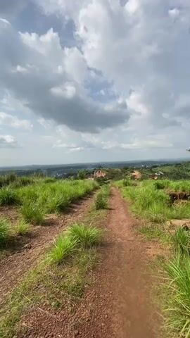 This is a POV video of someone walking up a dirt path on a grassy hill. The path is flanked by tall green grass on either side. In the distance, there is a view of a town or city with some buildings and trees. The sky is cloudy with patches of blue. As the person walks further up the hill, the view opens up to reveal a more expansive landscape. There is a noticeable cliff face on the right side of the path, with some trees growing on top of it. The path continues to ascend, leading towards the horizon.