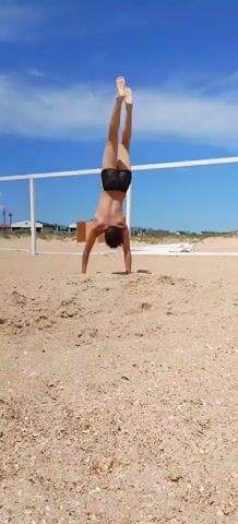 A person wearing black swim trunks is performing a gymnastic exercise on a sandy beach. They begin in a handstand with their legs extended upwards. Then, they lower their body into a bridge pose, with their hands and feet on the sand, and their back arched. The background shows a blue sky with white clouds, a white fence, and some distant buildings and sand dunes. The video consists of a few still frames, suggesting a slow-motion or stop-motion effect.