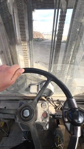 The video shows the first-person perspective from inside a forklift or similar heavy machinery. The driver's hand is visible on the steering wheel, and the dusty windshield offers a view of an outdoor industrial area. Through the forklift's lift mechanism, we see a person walking in the distance, a pile of wood or logs, and a building with a corrugated metal fence. The interior of the forklift appears worn and functional, with visible gauges and levers. The overall impression is of a work setting.