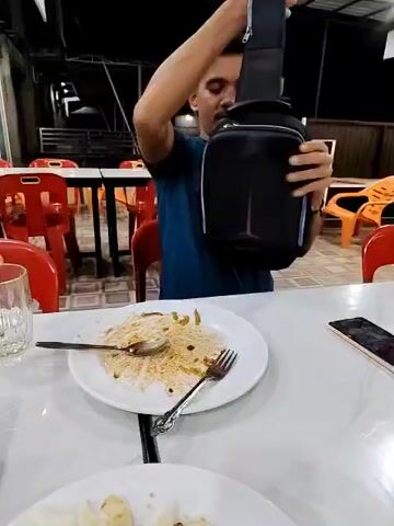 A man sits at a table in an outdoor restaurant. The table is white with a plate of leftover noodles and a cell phone on it.  He puts on a small black shoulder bag and reaches inside. The man is wearing a blue t-shirt with short sleeves. There are red plastic chairs surrounding the table. In the background are more tables and chairs, and a metal structure that appears to be part of the restaurant’s roof.