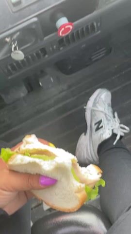 The video showcases the interior of a heavy machinery vehicle, likely a forklift or similar industrial vehicle. In the initial shot, a hand holding a half-eaten sandwich rests next to a foot wearing a white and silver New Balance sneaker. The dashboard displays various buttons and controls, including a prominent red emergency stop button. The view then shifts to the driver's perspective, revealing the steering wheel and instrument panel. The front windshield is cracked and has a sign taped to it. The setting appears to be an industrial yard with stacks of materials visible through the cracked windshield. The video seems to capture a moment of downtime or a break within the operational environment of the machinery.