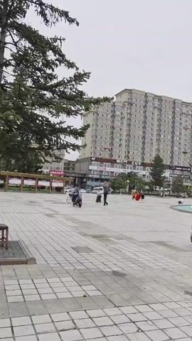 A wide shot shows an outdoor public space paved with square tiles. In the foreground, a parked electric scooter is visible on the right. In the middle ground, people are walking and children are playing. Further in the background, there are several multi-story buildings, trees, and a lamppost. The sky is overcast. The video pans slightly to the right.