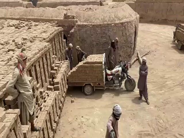 The video shows a brickyard with several men in traditional clothing. One man is standing near a tall stack of bricks, leaning against them. A three-wheeled vehicle, loaded high with bricks, is parked nearby. Several other men are standing around the vehicle, possibly loading or unloading the bricks. In the background, there are some circular brick structures, possibly kilns. The ground is sandy and dusty. The overall scene appears to depict manual labor in a hot, arid environment.