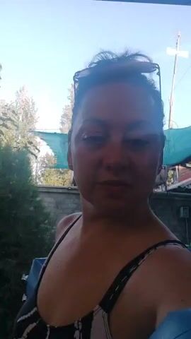 The video shows a woman with dark hair and sunglasses on top of her head. She is wearing a black and white patterned swimsuit and has a blue object partially visible on her right side. The background appears to be an outdoor setting with green foliage and a fence. The woman is looking directly at the camera.