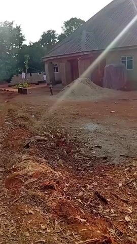 The video shows a dirt yard in front of a house. A pile of sand sits near the building. A person is seen walking in the yard. The camera moves to the right, passing by some banana trees and showing a dirt road littered with leaves and debris. There are other trees and buildings visible in the background.
