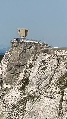 The video shows a rocky mountain with a military radar installation on its peak. The radar dish is large and rectangular, mounted on a structure that appears to be a building. The mountain itself is steep and rugged, with visible rock formations. The sky above is clear and blue. Two circular white objects are visible on the side of the mountain below the radar.
