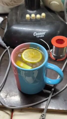 A close-up, slightly blurry shot focuses on a mug of tea with a slice of lemon and a piece of what appears to be ginger or lemongrass. The mug has a pink and blue design. In the background, out of focus, is a black electronic device labeled "Cosmos 12-LDA" and a spool of solder. The scene appears to be set on a wooden surface with some wires and cables visible.