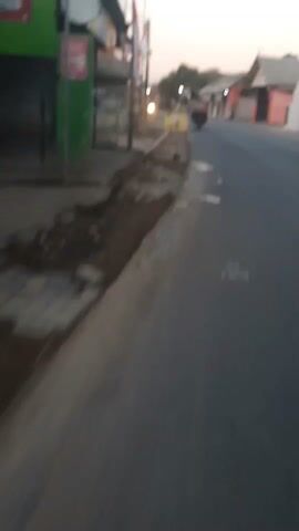 The video is a first-person perspective of someone riding a vehicle down a street. The street is paved and appears to be a two-lane road. On the left side of the street, there are buildings and shops. Some of the buildings are made of wood and appear to be old. There are also some trees and vegetation along the side of the road. On the right side of the street, there are more buildings, which appear to be houses. The sky is clear and it looks like it is either early morning or late evening. There are a few other vehicles on the road, including a motorcycle in the distance. The video is slightly blurry, which suggests that the person recording it is moving at a moderate speed. The overall scene is of a typical street in a rural or semi-urban area.