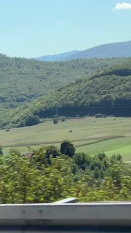 A video captures a view from a moving vehicle. The scene is dominated by lush green hills and mountains covered in dense trees. In the foreground, the tops of houses with red-tiled roofs are visible, nestled within a small village. A green road sign with white lettering indicating "Reskovici 339 m" is briefly in focus. The sky is a clear blue with scattered white clouds. The perspective suggests the viewer is looking out from a car or bus, with a railing of some kind visible at the bottom of the frame. The overall impression is a scenic, rural landscape on a sunny day.
