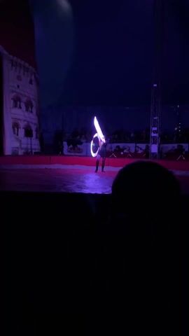 A person is performing on stage in a circus tent. They are wearing a short black outfit and are spinning two lit hula hoops. The lights on the hoops change color. The stage is lit in the same color as the hoops. There is an audience seated in the background. The performance is occurring at night or in a very dark space.