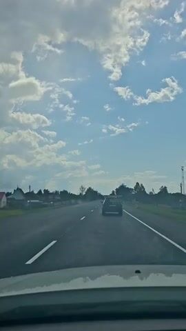 The video shows a view from inside a car traveling on a paved road. The sky is blue with scattered white clouds. Trees and buildings are visible on either side of the road. Another car is driving ahead of the camera car. The road has white lines marking the lanes.