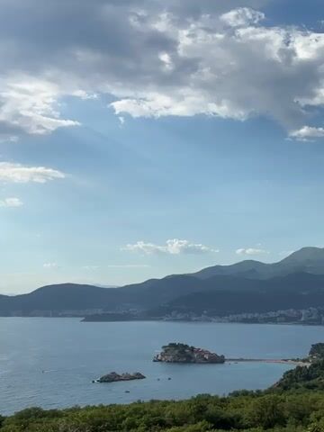 A wide-angle, vertical video shot from a high vantage point overlooking a coastal landscape. The scene is dominated by a vast expanse of blue water, meeting a shoreline lined with mountains in the distance. Sunlight filters through scattered clouds in the sky, creating visible sunbeams. In the middle ground, a small, rocky island is connected to the mainland by a narrow land bridge. The foreground consists of lush green foliage, suggesting the viewpoint is from a forested hillside. The overall impression is a serene, natural environment under a dynamic sky. There are no people or specific actions depicted.