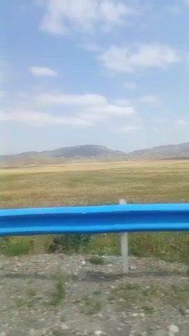 The video shows a view from a moving vehicle, looking out over a grassy field with rolling hills in the distance under a partly cloudy sky. A bright blue guardrail runs horizontally across the foreground, with white posts supporting it. The ground in the immediate foreground appears to be gravel and sparse vegetation. The video is short, lasting only a few seconds, and the camera movement suggests travel along a road.