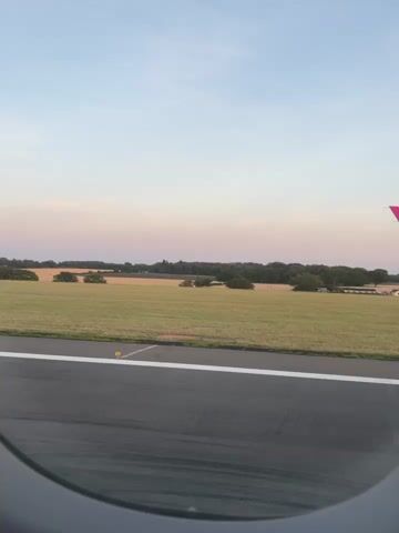 The video shows an airplane moving on a tarmac with a field and trees in the background. The sky is clear and shows the soft colors of either sunrise or sunset. The wing of the airplane is visible on the right side of the frame and has the Wizz Air logo on it. The image is taken from inside the airplane, looking out of a circular window. The tarmac is dark and appears wet, with white markings indicating a runway.