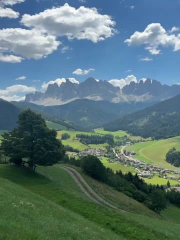A wide shot captures a scenic view of a valley with a village nestled within it, surrounded by lush green hills and dramatic mountain peaks under a partly cloudy sky. In the foreground, a grassy slope dominates the left side of the frame, with a dirt path winding its way down. A large, dark green tree stands prominently on the left side of the slope, its branches casting shadows. The middle ground reveals a charming village with red-roofed houses clustered along a road, interspersed with fields and smaller trees. The background is dominated by towering, jagged mountain ranges, their peaks sharp against the blue sky dotted with white clouds. The overall scene conveys a sense of peacefulness and natural beauty. There are no people or animals visible in the video.