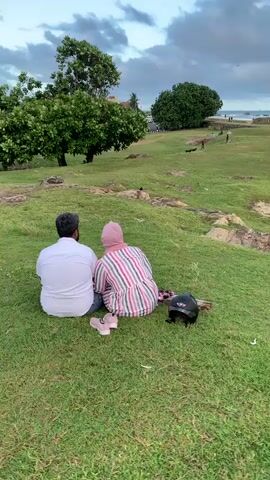 This media file captures a serene outdoor scene in Galle, Sri Lanka. In the foreground, a couple sits on a grassy incline, their backs to the camera, enjoying the view. The man wears a white shirt and jeans, while the woman is dressed in a striped outfit and a pink hijab. A black helmet and a pair of pink sneakers are placed beside them on the grass.

The background unfolds into a wide, open field with more people scattered across it, some walking, some appearing to be playing with dogs. Beyond the field, the ocean stretches out, with waves crashing against a coastal structure, possibly a fort or old wall. The sky above is partly cloudy, suggesting it might be late afternoon or early evening. The overall atmosphere appears peaceful and relaxed, with the couple seemingly enjoying a quiet moment together amidst the gentle activity of the location. No visible text is present in the media.