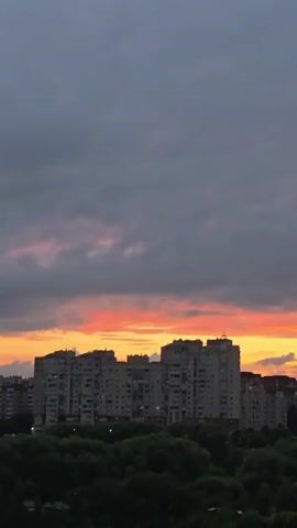 This is a vertical, slow-motion video of a sunset over a city. The sky is filled with clouds, with the bottom portion illuminated by the setting sun in shades of orange and pink. Below the sky, there is a line of apartment buildings, followed by a dense band of green trees. The foreground shows a dark, blurry area, likely representing the ground or a lower perspective. The overall mood is peaceful and serene.