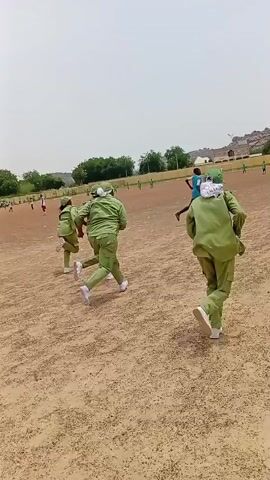 A group of people in matching green uniforms are running across a dusty, open field under a cloudy sky. The field appears to be a sports field, with some green markings visible in the distance and trees lining the background. The individuals are running in a group, some spread out more than others. The camera is positioned low to the ground, following the runners. Buildings and a hillside are visible in the background.