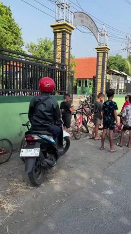 The video shows a group of young boys gathered outside a school gate. One boy in a red shirt is sitting on a red bicycle and appears to be shouting or laughing. Another boy, wearing a black t-shirt, stands near a bicycle and holds a bottle of liquid. Two other boys are visible in the background, one in a white t-shirt with a palm tree design and another boy wearing a dark blue and black patterned outfit. There are several bicycles and a motorcycle in the scene, and the environment appears to be an outdoor area, possibly a street or courtyard. The gate features decorative pillars with yellow and black patterns.