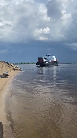 A ferry boat navigates a wide, calm river under a dramatic sky filled with dark, heavy clouds that suggest an impending storm. The ferry, a white and blue vessel with a deck for vehicles, is moving away from the camera towards the horizon. On the left side of the frame, a sandy shore is visible, with some debris and rocks. The water is a murky brown, reflecting the overcast sky. In the distance, a treeline marks the far bank of the river. The overall mood is somber and a bit foreboding, amplified by the contrast between the dark clouds and the brighter, sunlit patches in the distance. The time of day appears to be late afternoon or early evening. The location is identified as Selishche Govyadino, Russia. There is some Cyrillic text visible on the side of the ferry, but it is too small to be read clearly.