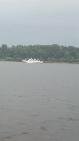 The video shows a wide, calm river with a wooded shoreline in the background. A white ship with red accents is anchored near the shore. The sky is overcast and gray.