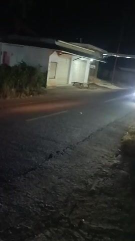 This is a night time video of a street scene. A car approaches and its headlights illuminate a small building with what appears to be a covered patio or porch. Two red lights on the back of the car are visible as it passes. A motorcycle with two riders is parked in front of the building.
