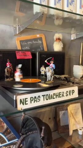 The video shows a close-up of a shelf in what appears to be a shop or display area. On the shelf, there are several objects including a record player with a vinyl record on it. On the record, there are small figurines of people on horses. There are also two speakers behind the record player, and a small chalkboard with writing on it. A fluffy toy is on top of one of the speakers. To the right of the speakers, there is a white object that looks like a roll of toilet paper or paper towels. In the foreground, there is a sign that says "NE PAS TOUCHER SUP". Below the shelf, there are more items on display, including what looks like a hat and some papers. The background of the video is a blurry view of the shop interior, with shelves filled with various items. The lighting suggests an indoor environment.