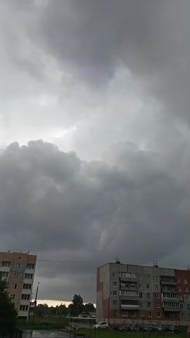 The video shows a slightly tilted view of a cloudy sky over two apartment buildings. The sky is filled with dark, ominous clouds, suggesting an approaching storm. The buildings appear to be multi-story residential structures. The lower parts of the buildings show parked cars and some green space. The overall atmosphere is somber due to the heavy cloud cover.