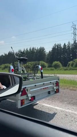 The video shows a person driving a car. The car is towing a trailer on which a white and green tricycle is loaded. The tricycle has a basket on the front. The car is black and appears to be a sedan. The driver is visible in the side mirror. The scene is set on a sunny day, with trees and power lines visible in the background. The road appears to be paved.