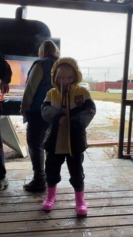 A young child wearing a yellow and black hooded jacket, black pants, and pink rubber boots dances on a wooden porch. The child is facing the camera and makes exaggerated movements, raising arms and swaying back and forth. Behind the child, two adults stand near a smoking grill. One adult wears a blue vest and the other is partially obscured. The porch has railings and is wet, with puddles visible. In the background, there is a yard with patches of snow, a house, and a car. The overall setting appears to be a backyard barbecue on a damp or early spring day.