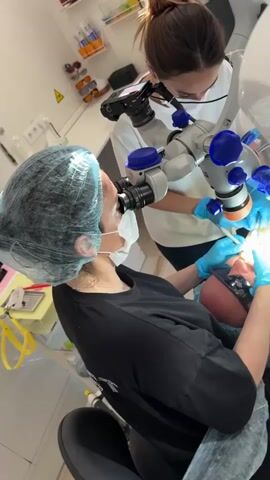 A dental professional in a black shirt and blue gloves, wearing a hairnet and mask, looks into a dental microscope. The microscope is positioned above a patient who is lying down with protective eyewear. Another dental professional, wearing a white top, is assisting. The microscope has several blue adjustment knobs and an orange component. A dental instrument is being used in the patient's mouth. The scene appears to be a dental operatory with medical equipment visible in the background.
