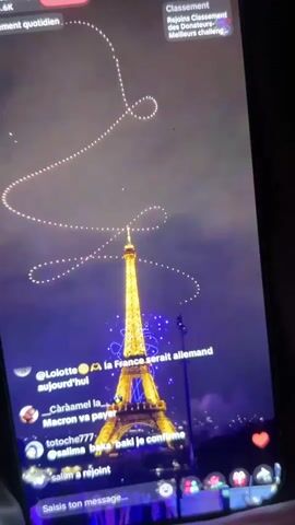 The video shows a nighttime view of the Eiffel Tower in Paris, illuminated with lights. A drone or light show is creating patterns in the sky above the tower. There are comments overlaid on the video, with some users expressing political opinions. In the lower right corner, people are visible, some holding phones and one person appears to be taking a selfie. The overall impression is of a live stream of a nighttime event in Paris.