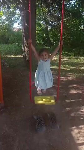 A young girl in a light blue dress is on a swing set. She is holding onto the red ropes of the swing. The swing seat is yellow. There are two tires on the ground below the swing. The girl is smiling at the camera. The background is a park with trees and grass. The sun is shining.
