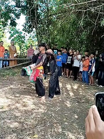 The video captures an outdoor scene where two individuals in dark attire are performing a choreographed martial arts display in front of a crowd. The performers are wearing traditional garb, and the performance appears to involve intricate movements and simulated combat techniques. A third person is also participating wearing striped shirt and white pants. The crowd is composed of men, women, and children, who are watching the performance with interest. The setting is under the shade of trees, with a dirt ground.  A small wooden fence partially separates the audience from the performance area.