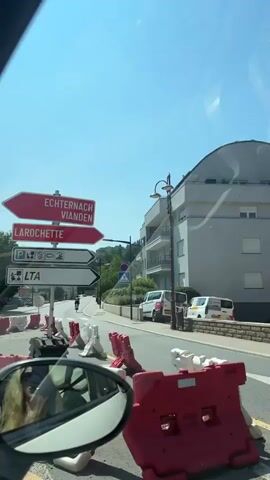 The video begins with a shot from inside a car, looking out the windshield and side mirror. Ahead, a road is lined with red and white construction barriers. Traffic signs indicate directions to "ECHTERNACH," "VIANDEN," and "LAROCHETTE." A pedestrian crossing is visible, and a cyclist is in the distance. The car moves forward, passing the barriers. The view then shifts to the road ahead, showing houses on either side and parked cars. A silver car is in front, also traveling on the road. The video continues to follow the road as it curves slightly, with more houses and trees visible. The sky is clear and blue. The car appears to be in a town or village.
