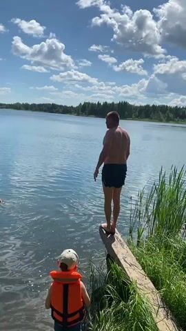A man dives into a lake from a wooden plank. A child wearing an orange life vest watches from the shore. Another person is already in the water to the left. The sky is blue with white clouds, and trees line the opposite shore.