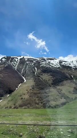 A vehicle is moving on a road on a sunny day. On the left side of the road is a mountainside with snow visible near the top. In the foreground, a guard rail is visible. The sky is blue and partly cloudy.  As the vehicle moves, the mountain appears to rotate through the window.