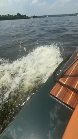The video begins with a close-up of a dark green boat's bow, showing a wooden deck and a black rope. White water splashes up from the wake. The camera pans slightly to the right, revealing more of the boat's interior and the water. In the distance, several sailboats with colorful sails are visible on a wide body of water, with a tree-lined shore in the background under a partly cloudy sky. The boat is moving forward, creating a significant wake. The scene suggests a leisurely day on the water, possibly during a sailing regatta.